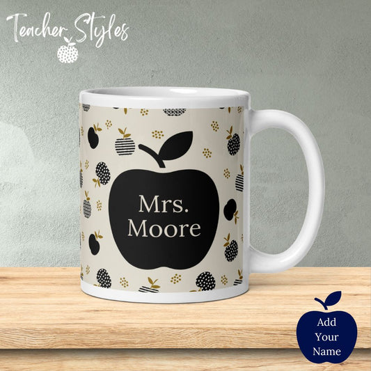 Our lovely white ceramic teacher mug is covered with sophisticated taupe background covered in pattern of black apples made of solids, stripes and dots. Gold-brown leaf accents add a bit of pop. Each side of the mug features a large black apple that we can personalize with your name!  Mug on table .