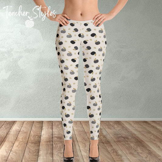 Modern Apple Leggings by Teacher Styles. Pattern has off-white background with black apples in solids, stripes and dots. Goldish-brown leaf accents. Model standing in room.