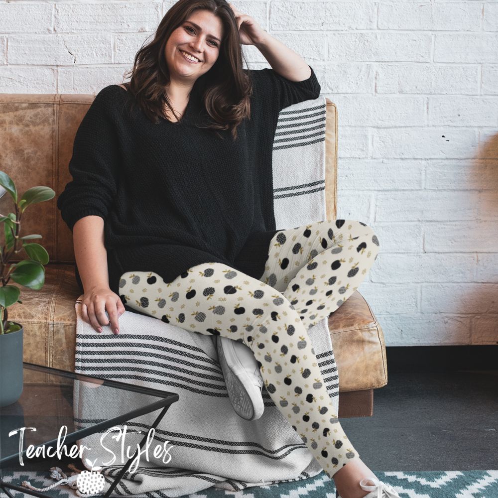 Modern Apple Plus Size Leggings by Teacher Styles. Image of happy woman sitting on sofa wearing modern apple leggings and black over-sized shirt. Light beige/cream leggings feature a pattern of black apples in solids, stripes and dots.  